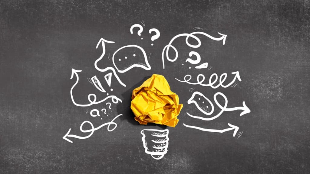 Crumpled yellow paper shaped like a lightbulb surrounded by white doodles on a chalkboard background